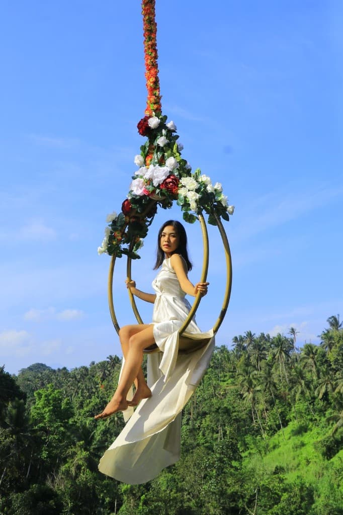 A guest in a white dress on the flower-draped gold libra swing above the jungle at Aloha Ubud Swing